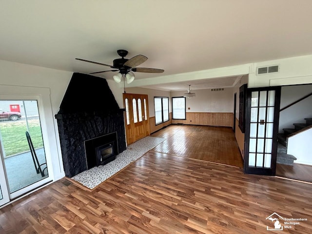 unfurnished living room with wood finished floors, stairs, a wainscoted wall, ceiling fan, and visible vents