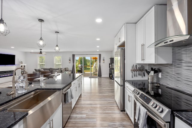 kitchen featuring wall chimney range hood, appliances with stainless steel finishes, hanging light fixtures, white cabinets, and dark stone counters