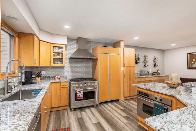 kitchen with a wood stove, premium appliances, a sink, wall chimney range hood, and light wood-type flooring