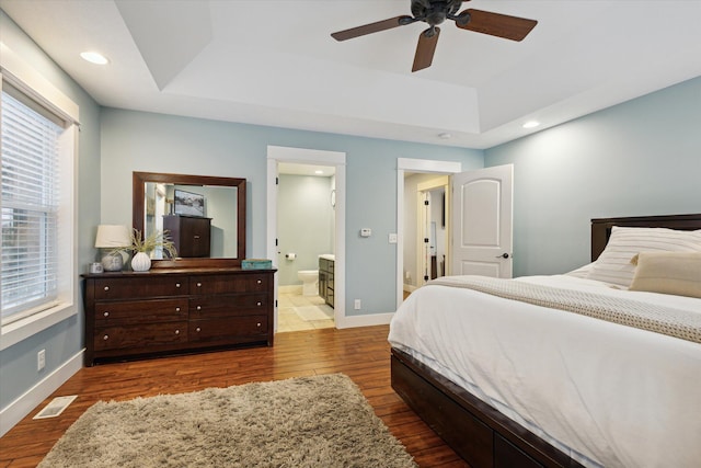 bedroom with hardwood / wood-style floors, connected bathroom, a raised ceiling, and ceiling fan