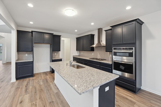 kitchen featuring a center island with sink, wall chimney range hood, sink, light wood-type flooring, and tasteful backsplash