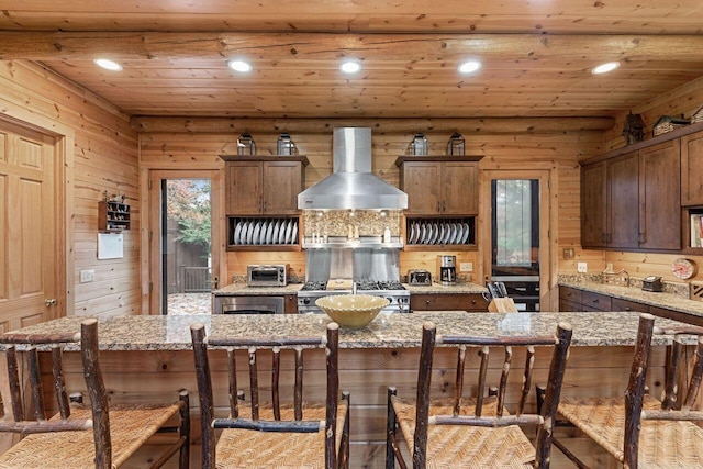 kitchen featuring stainless steel stove, wall chimney exhaust hood, a kitchen island, light stone counters, and a breakfast bar area