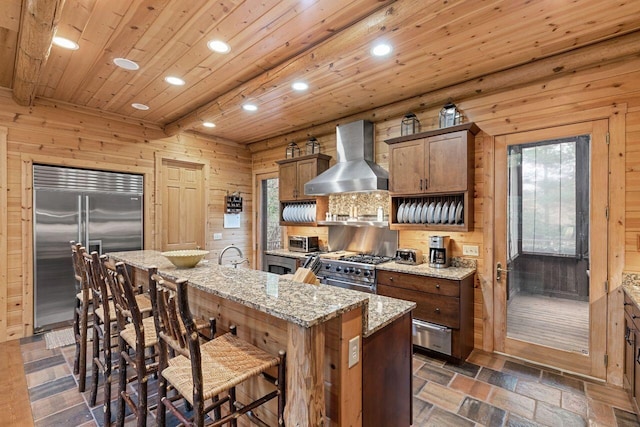 kitchen featuring high end appliances, wall chimney range hood, an island with sink, and a wealth of natural light