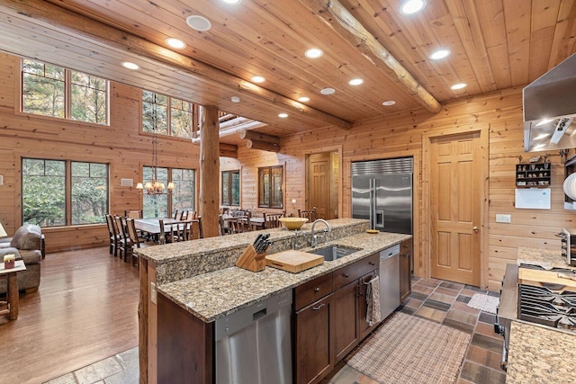 kitchen featuring a center island with sink, sink, wooden walls, beamed ceiling, and stainless steel appliances