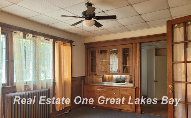 interior space with ceiling fan, radiator heating unit, wooden walls, and dark colored carpet