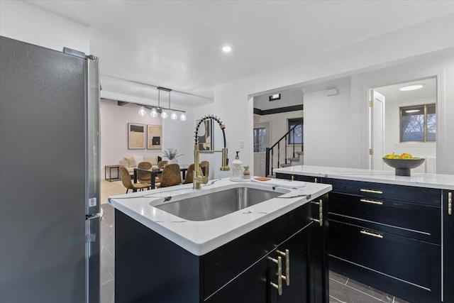 kitchen with a sink, light stone counters, freestanding refrigerator, dark cabinetry, and a kitchen island with sink