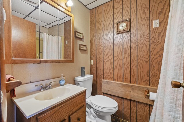 bathroom with a paneled ceiling, wood walls, vanity, and toilet