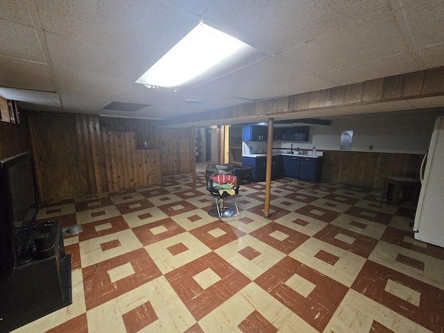 basement with a paneled ceiling, wood walls, and white refrigerator