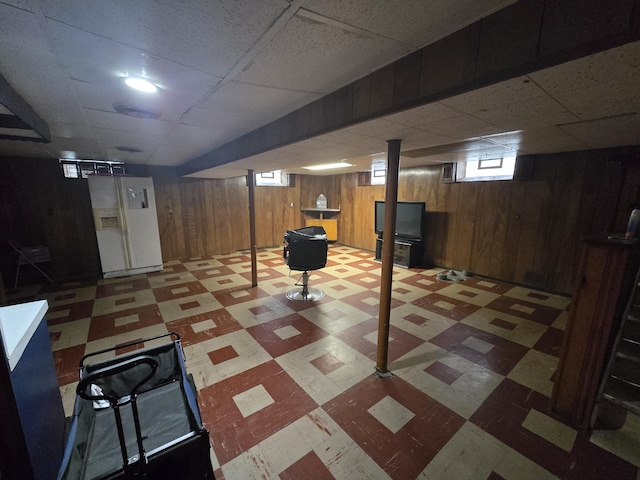 basement featuring a paneled ceiling, white fridge with ice dispenser, and wood walls