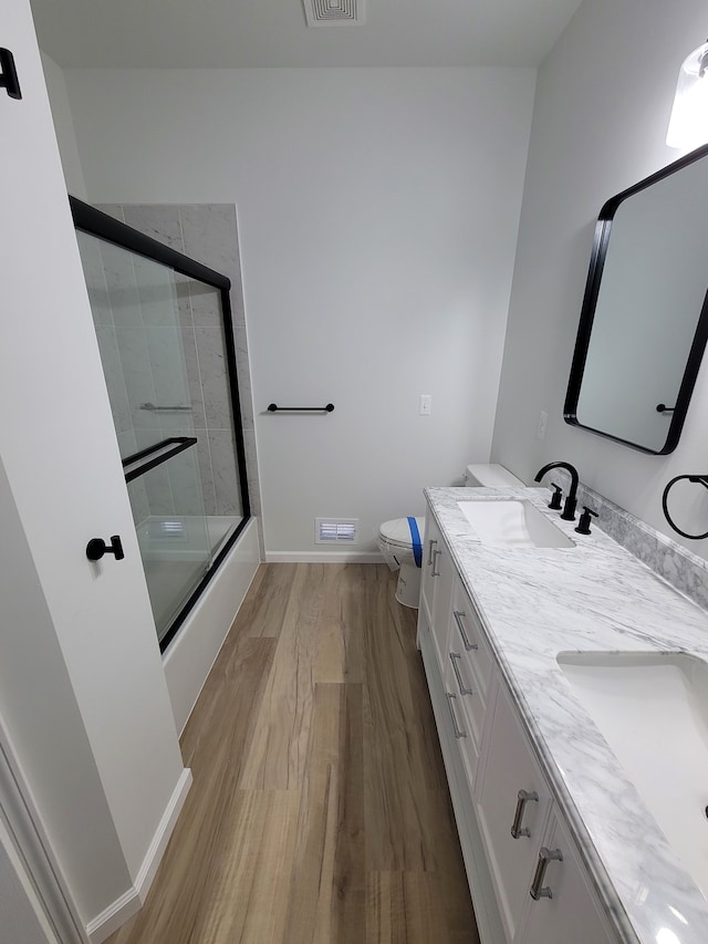 bathroom with double vanity, toilet, enclosed tub / shower combo, a sink, and wood finished floors