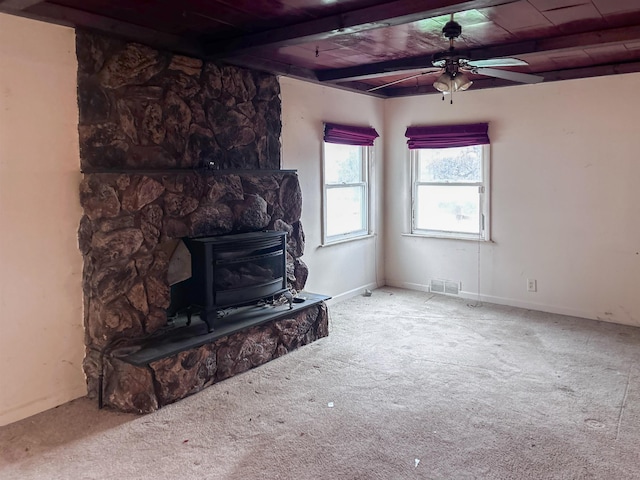 unfurnished living room featuring beamed ceiling, carpet floors, a wood stove, and ceiling fan