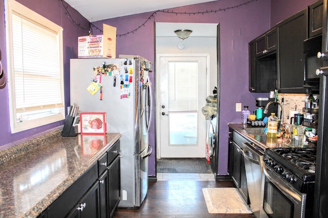 kitchen with appliances with stainless steel finishes, backsplash, dark wood-type flooring, dark stone countertops, and sink