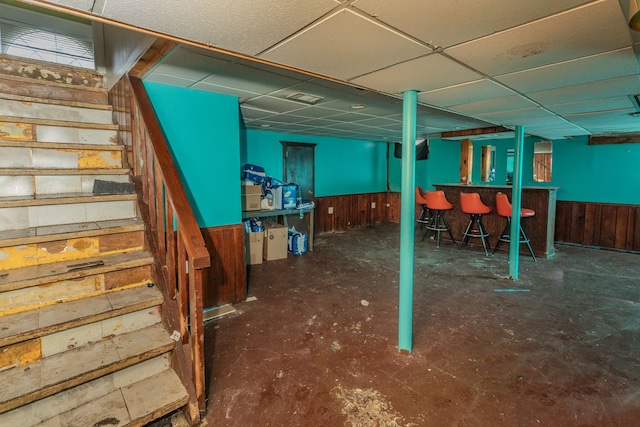 basement featuring a drop ceiling, wood walls, and bar area