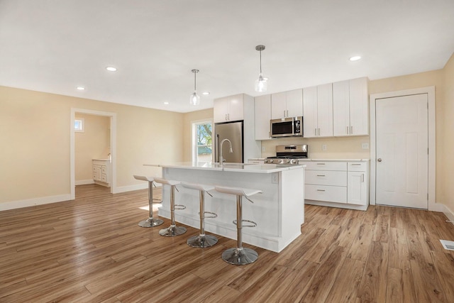 kitchen with appliances with stainless steel finishes, a kitchen island with sink, pendant lighting, light hardwood / wood-style floors, and white cabinetry