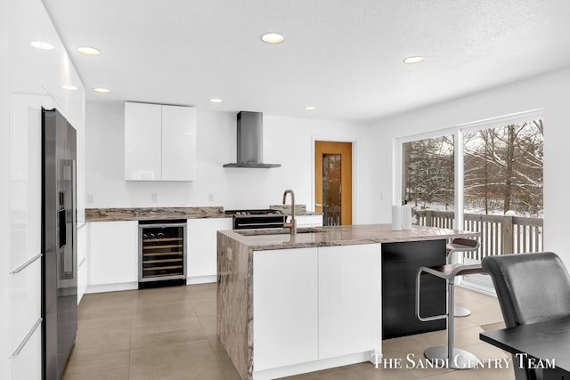 kitchen featuring a center island with sink, wall chimney exhaust hood, sink, white cabinetry, and wine cooler