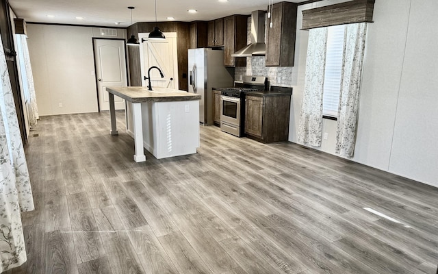 kitchen with appliances with stainless steel finishes, wall chimney range hood, pendant lighting, a center island with sink, and light hardwood / wood-style floors