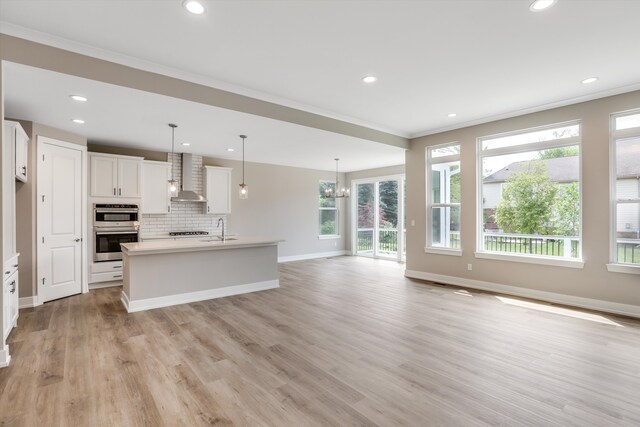 kitchen with a kitchen island with sink, wall chimney range hood, hanging light fixtures, light hardwood / wood-style flooring, and white cabinetry