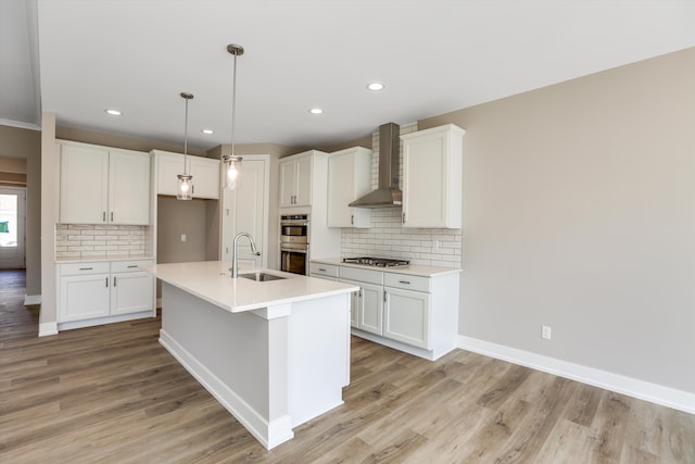 kitchen featuring an island with sink, light wood-type flooring, wall chimney range hood, and appliances with stainless steel finishes