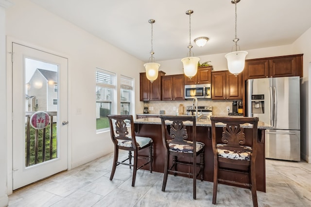 kitchen featuring light stone countertops, an island with sink, pendant lighting, decorative backsplash, and appliances with stainless steel finishes