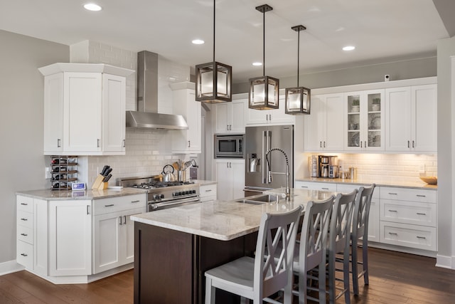 kitchen featuring white cabinets, wall chimney range hood, an island with sink, hanging light fixtures, and high quality appliances