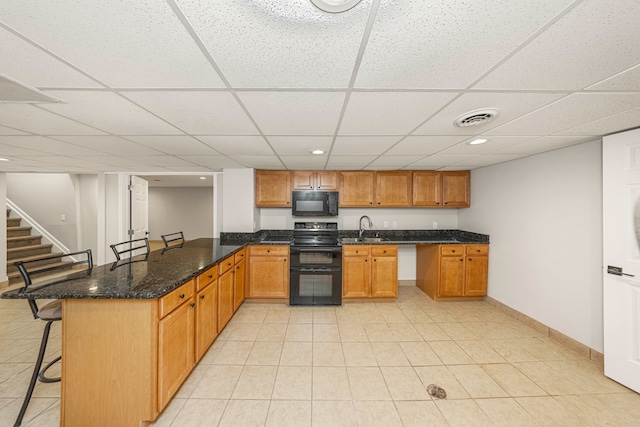 kitchen featuring a drop ceiling, dark stone counters, sink, black appliances, and a breakfast bar area