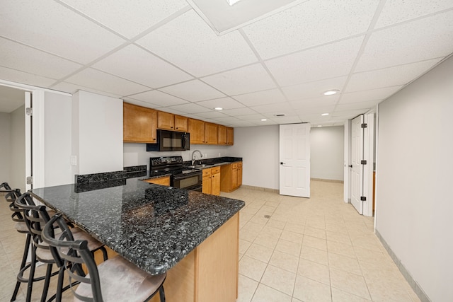 kitchen with a drop ceiling, dark stone counters, black appliances, a kitchen bar, and kitchen peninsula