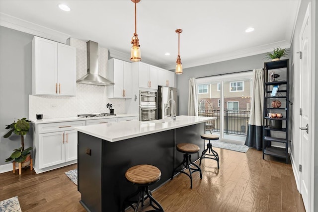 kitchen with stainless steel appliances, white cabinetry, wall chimney exhaust hood, pendant lighting, and a kitchen island with sink