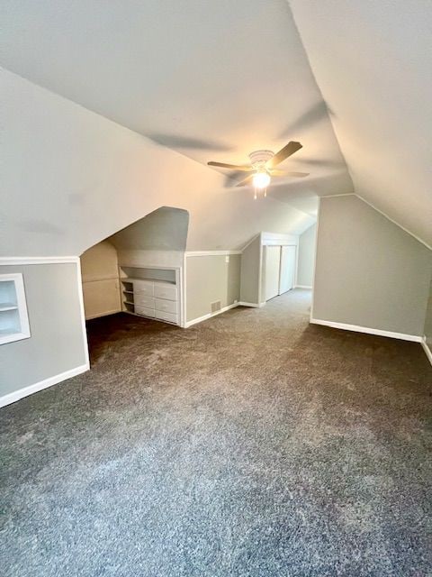 additional living space with dark colored carpet, built in shelves, vaulted ceiling, and ceiling fan