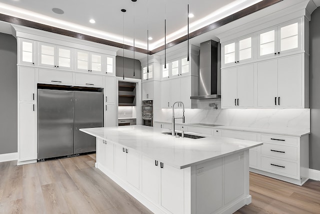 kitchen with a large island with sink, black appliances, wall chimney exhaust hood, decorative light fixtures, and light stone counters