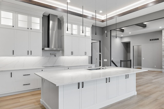 kitchen with sink, wall chimney exhaust hood, decorative light fixtures, a kitchen island with sink, and white cabinets