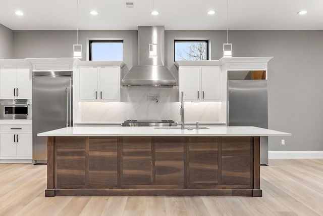 kitchen featuring stainless steel appliances, hanging light fixtures, wall chimney exhaust hood, and an island with sink