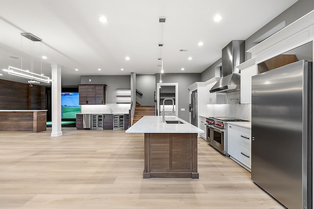 kitchen with sink, wall chimney exhaust hood, an island with sink, decorative light fixtures, and stainless steel appliances