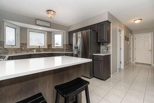 kitchen with light tile patterned floors, dark brown cabinets, a kitchen breakfast bar, stainless steel refrigerator with ice dispenser, and decorative backsplash