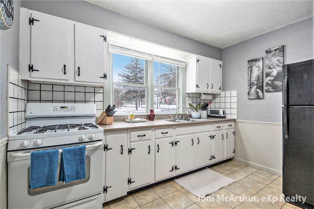 kitchen with a wainscoted wall, white range with gas cooktop, freestanding refrigerator, white cabinets, and a sink