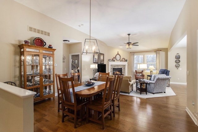 dining space featuring a fireplace, ceiling fan with notable chandelier, vaulted ceiling, and dark wood-type flooring