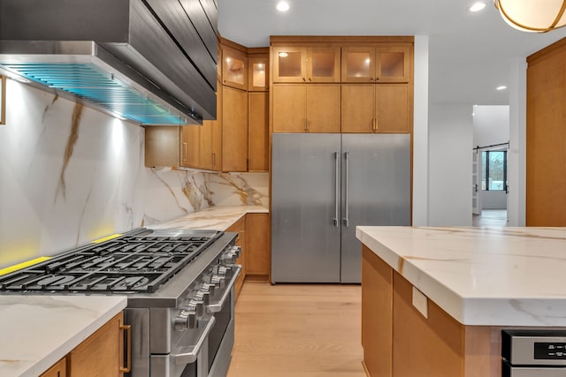 kitchen with backsplash, light stone counters, premium appliances, wall chimney range hood, and light hardwood / wood-style flooring