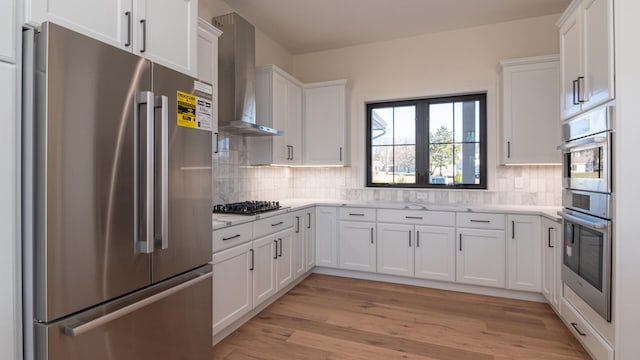 kitchen featuring appliances with stainless steel finishes, backsplash, wall chimney exhaust hood, light hardwood / wood-style floors, and white cabinetry