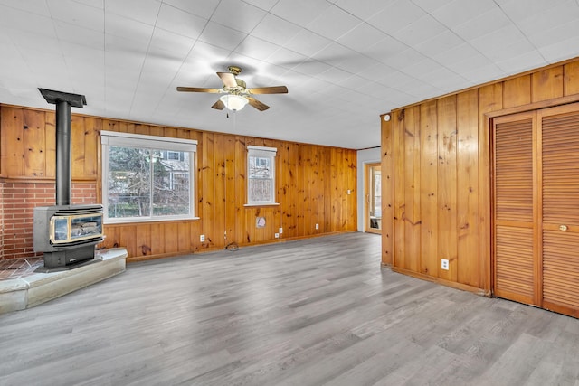 unfurnished living room with light wood-type flooring, a wood stove, ceiling fan, and wood walls