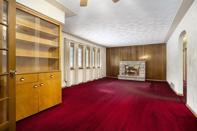 unfurnished living room featuring a fireplace, dark carpet, ceiling fan, and wood walls