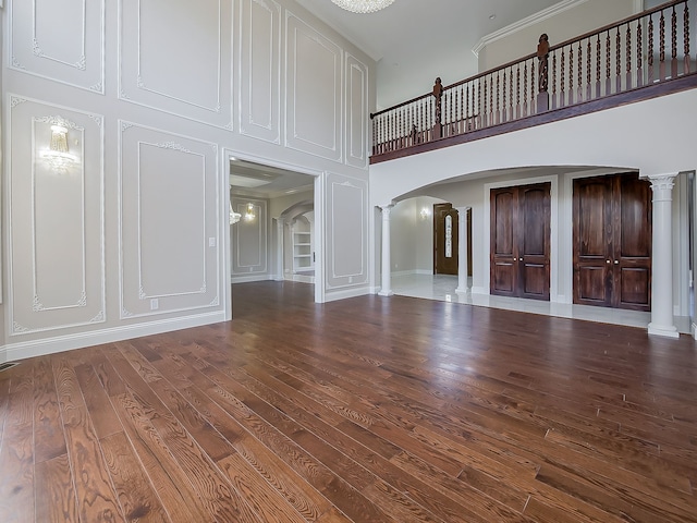 unfurnished living room featuring a towering ceiling, ornate columns, and dark wood-type flooring