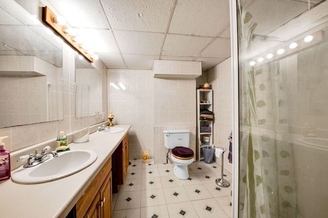 bathroom with a drop ceiling, an enclosed shower, toilet, vanity, and tile walls