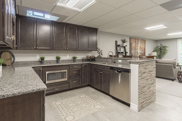 kitchen with kitchen peninsula, light stone countertops, appliances with stainless steel finishes, dark brown cabinets, and sink