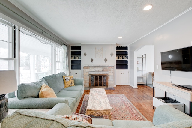 living room with a stone fireplace, crown molding, built in shelves, a textured ceiling, and light hardwood / wood-style floors