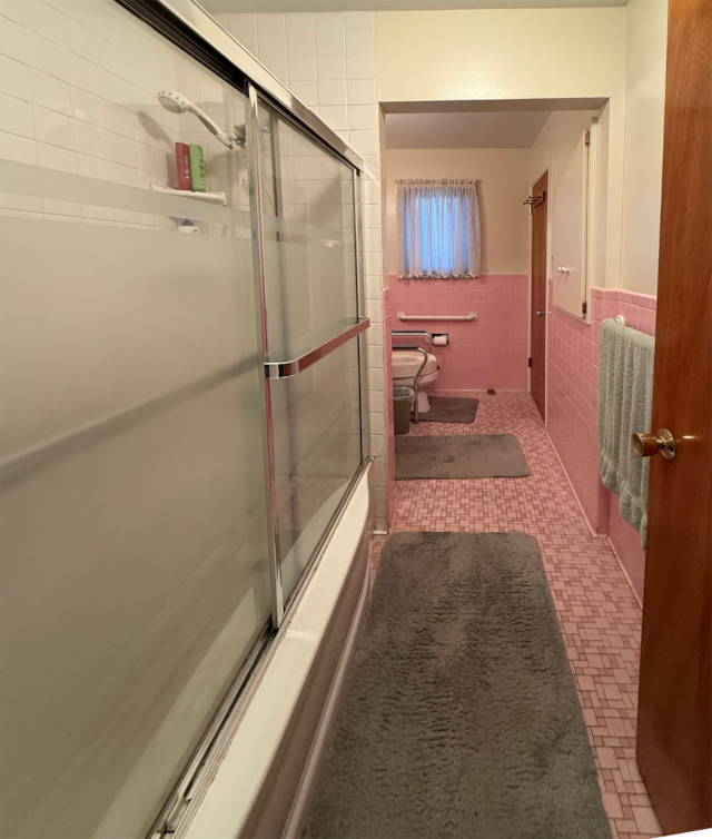 bathroom featuring toilet, combined bath / shower with glass door, wainscoting, and tile walls