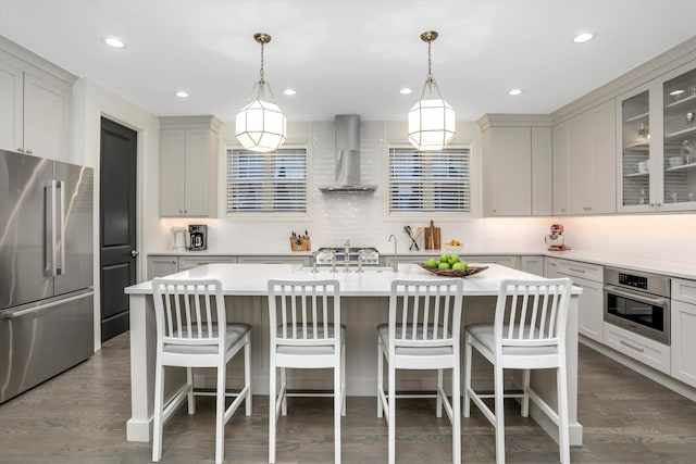 kitchen with wall chimney range hood, decorative light fixtures, a breakfast bar area, a kitchen island with sink, and appliances with stainless steel finishes