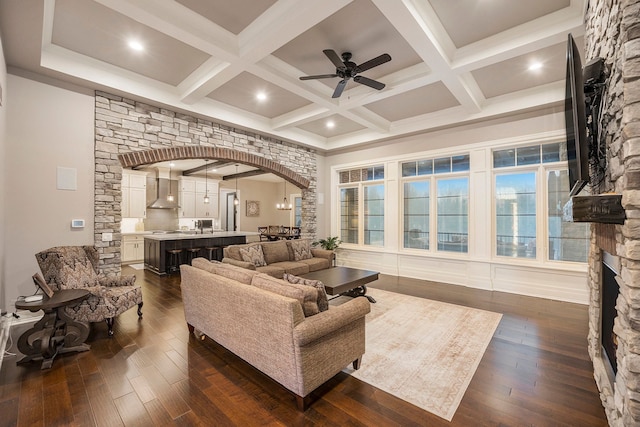 living room with dark wood-type flooring, coffered ceiling, a stone fireplace, ceiling fan, and beam ceiling