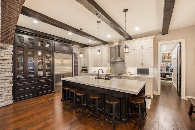 kitchen featuring wall chimney range hood, hanging light fixtures, appliances with stainless steel finishes, beam ceiling, and a kitchen bar