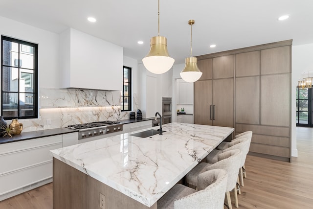 kitchen with hanging light fixtures, stainless steel gas cooktop, an island with sink, dark stone counters, and decorative backsplash