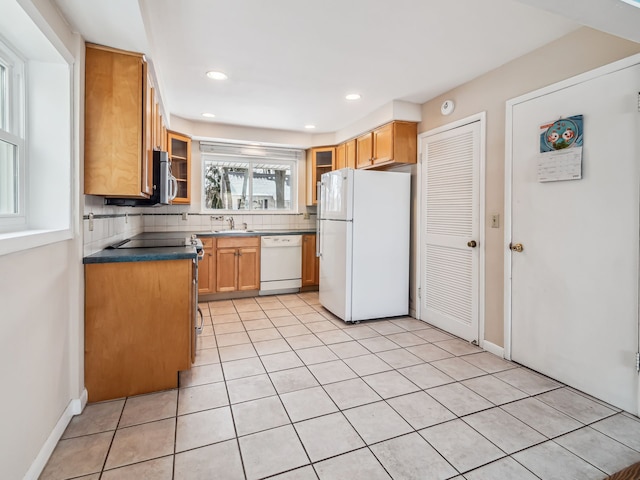 kitchen featuring tasteful backsplash, dark countertops, glass insert cabinets, brown cabinetry, and white appliances