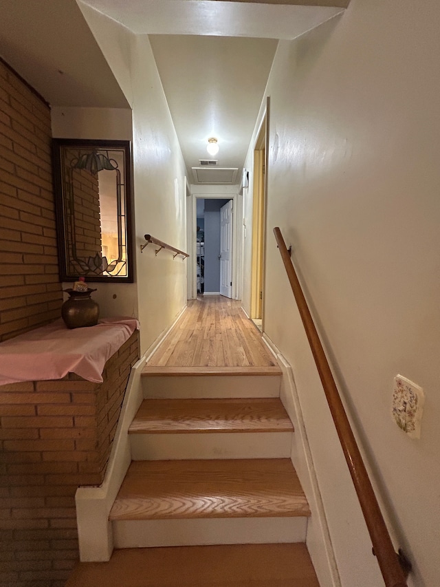 stairway with brick wall, attic access, and wood finished floors